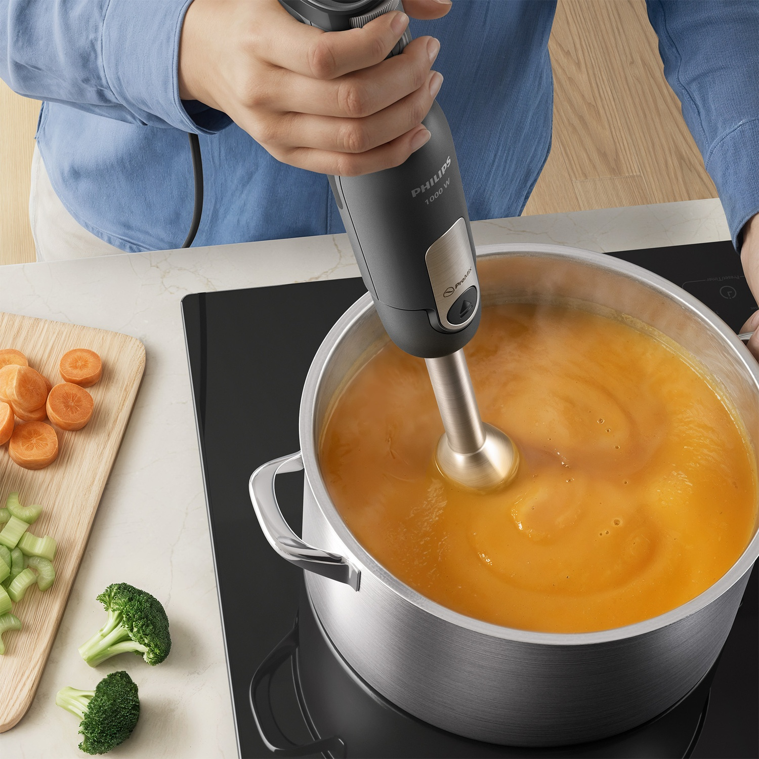 A person uses a grey Philips hand blender to puree orange soup in a pot on a stove next to sliced carrots and broccoli.