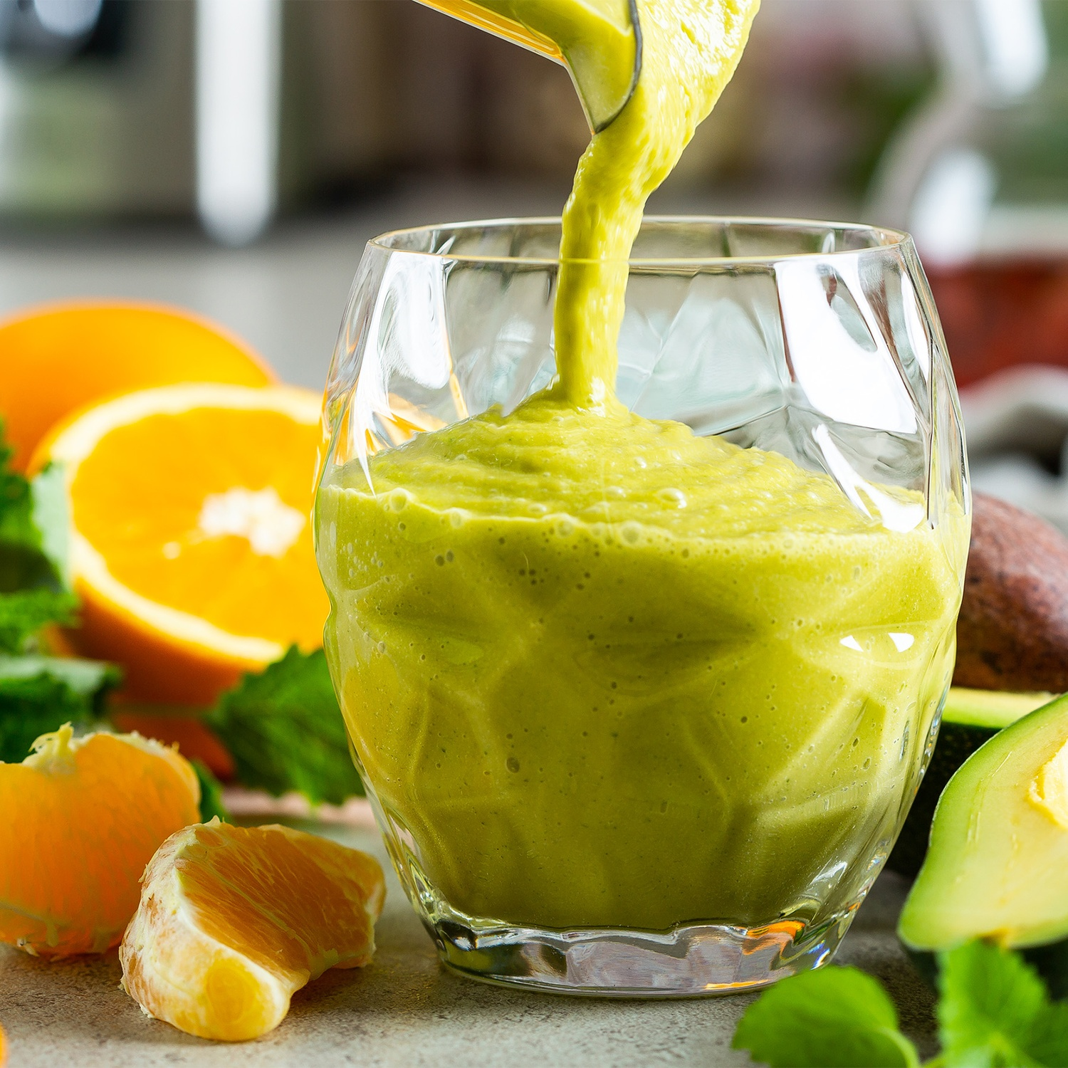 A thick green smoothie is poured into a faceted glass, surrounded by sliced oranges and avocado on a table.