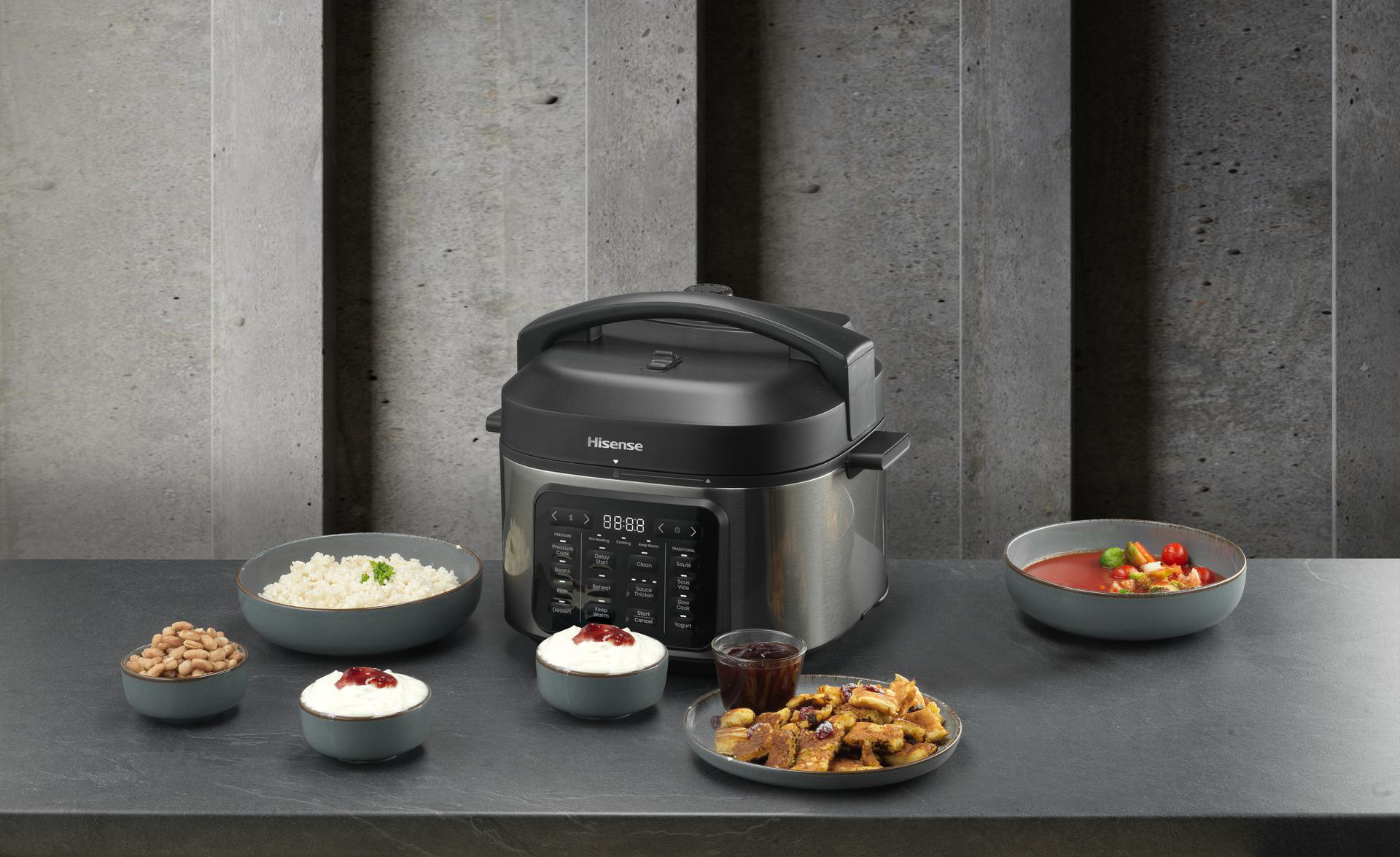 Ein Hisense-Multikocher auf einer grauen Theke, umgeben von Schüsseln mit Reis, Bohnen, Suppe, Joghurt und Pfannkuchen.