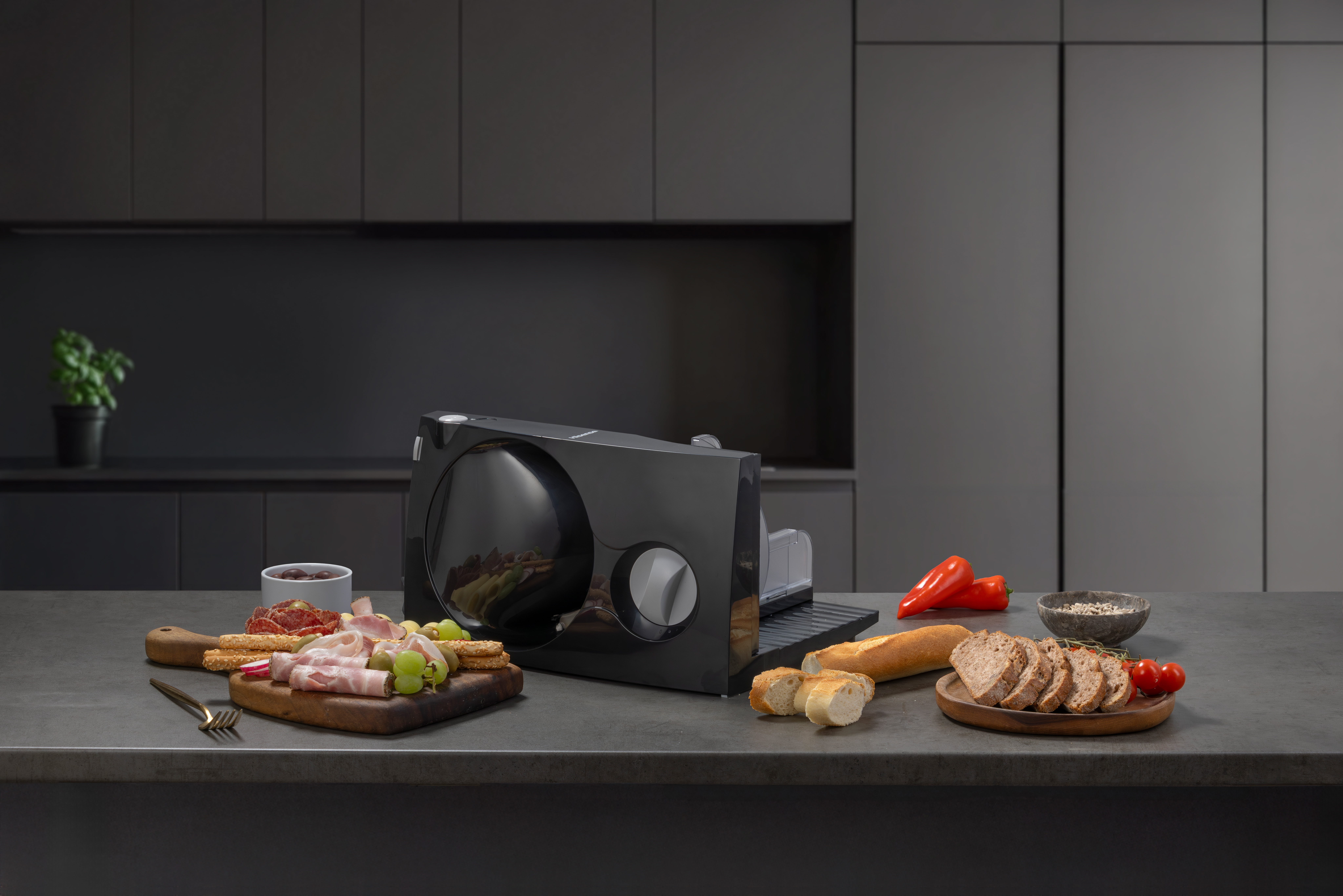 Eine schwarze Allesschneider-Maschine steht auf einer Küchentheke, umgeben von Wurst, Brot, Weintrauben und Paprika.