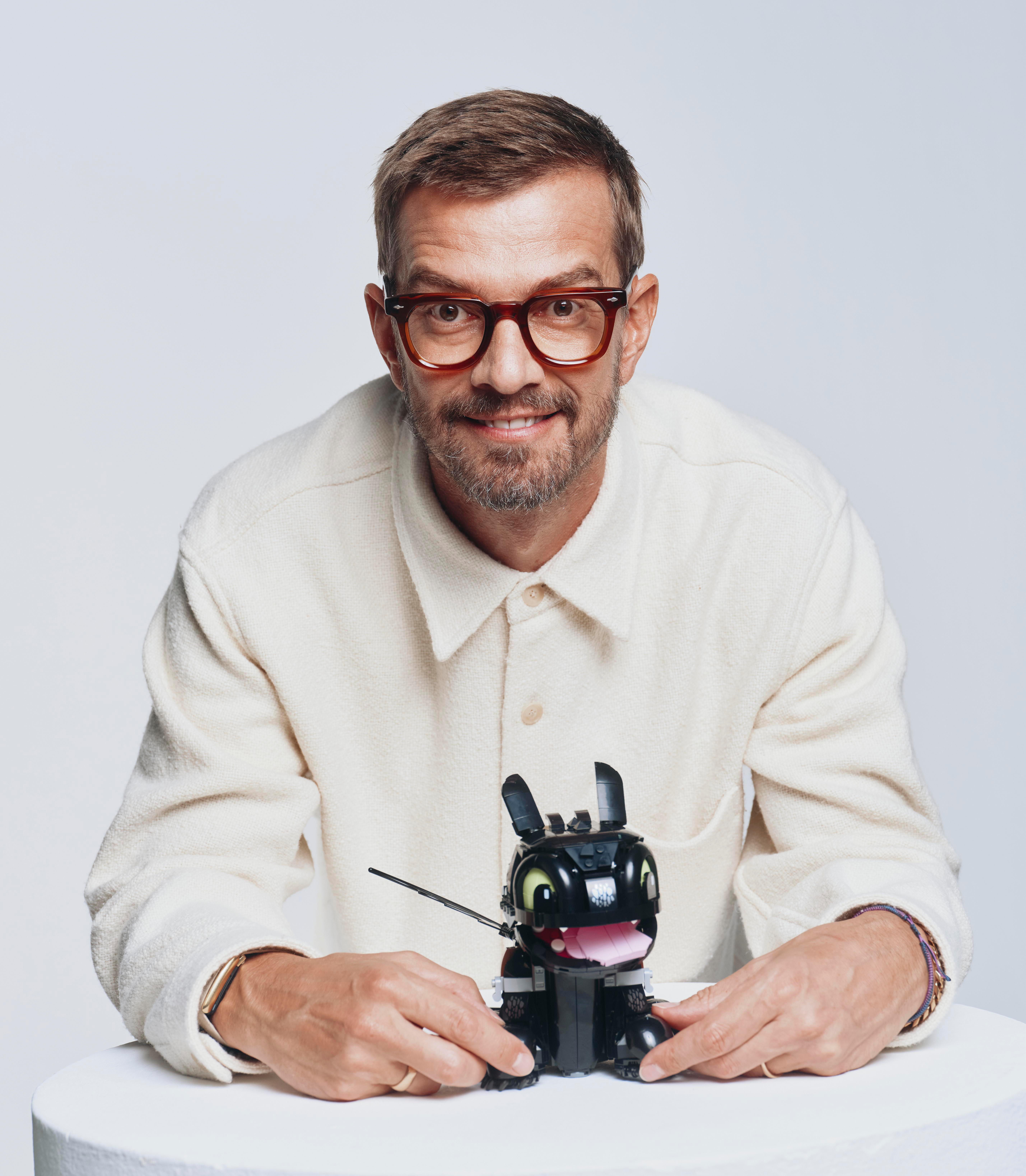 Ein lächelnder Mann mit Brille in einem weißen Hemd sitzt am Tisch und hält ein kleines schwarzes Spielzeugtier.