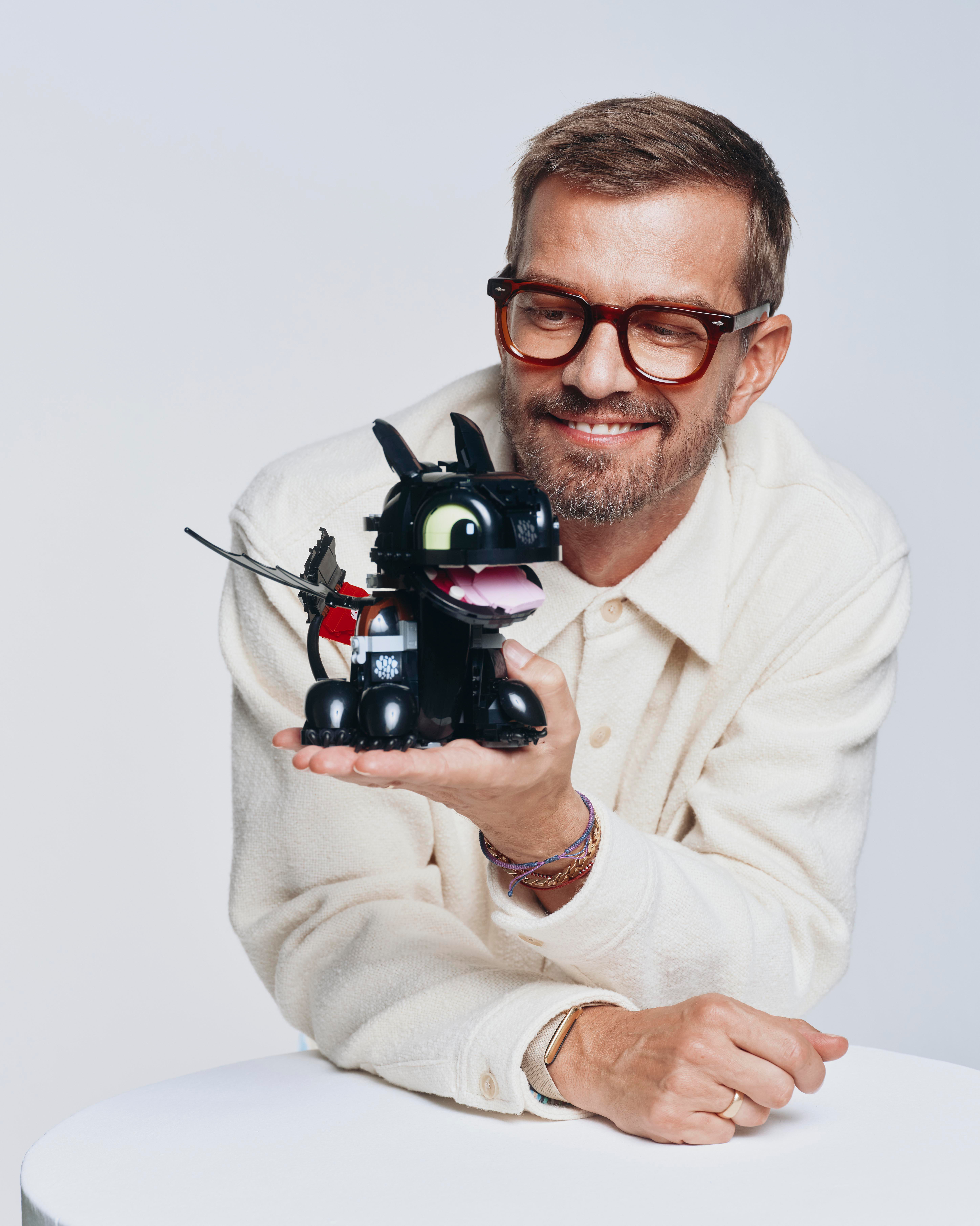 Ein lächelnder Mann in weißem Hemd und Brille hält einen kleinen schwarzen Legodrachen in den Händen.