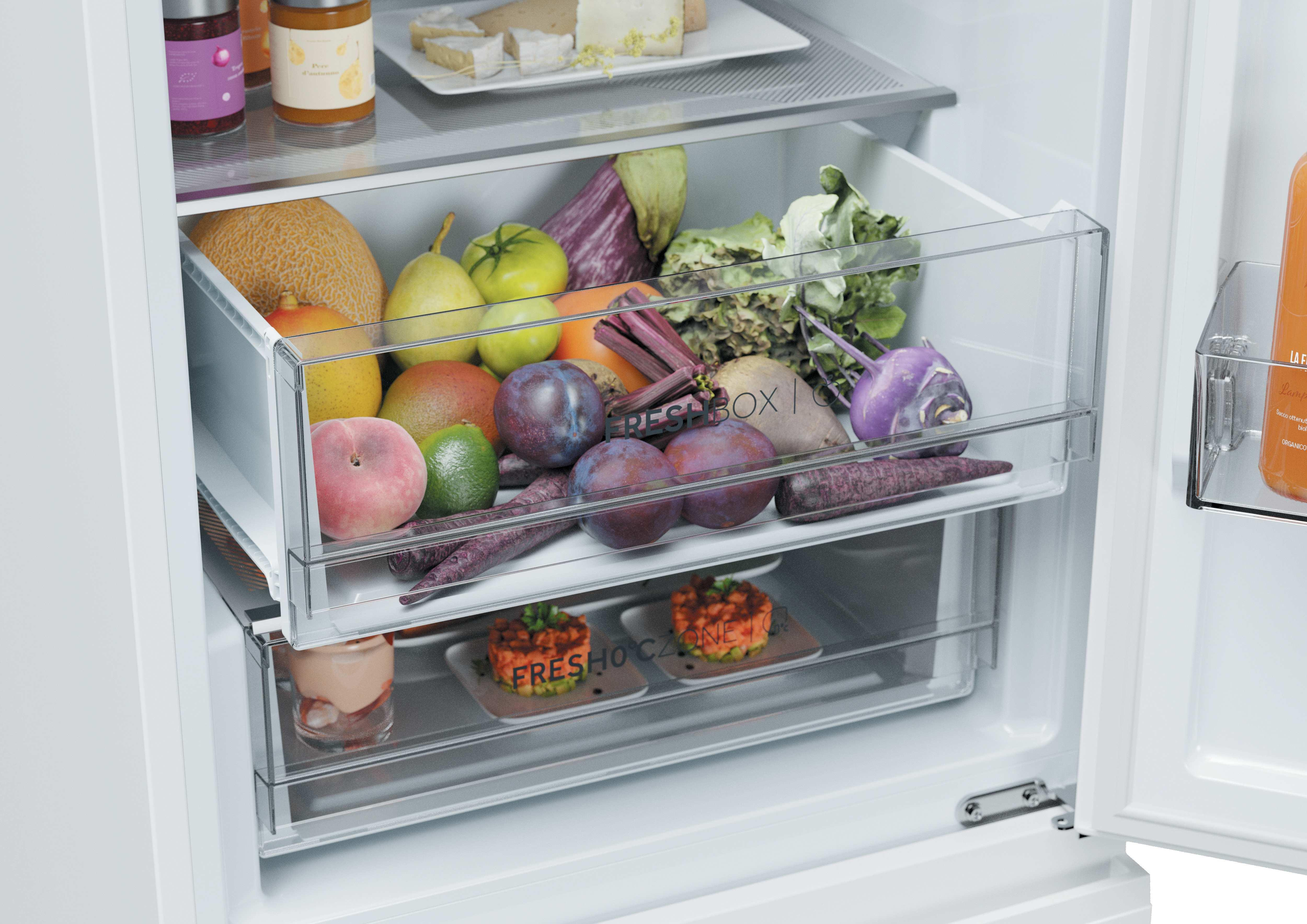 Un refrigerador abierto con un cajón lleno de frutas y verduras, y platos pequeños en el compartimento inferior.