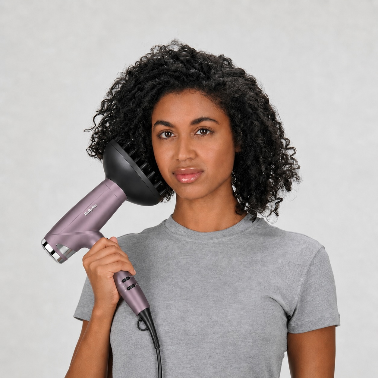 Eine Frau mit lockigem Haar hält einen Lavendelfarbener Föhn mit Diffusor.