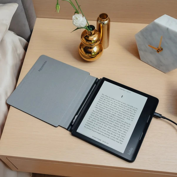 Otwarty czytnik e-booków z szarym etui i rysikiem leży na szafce nocnej z wazonem i zegarem.