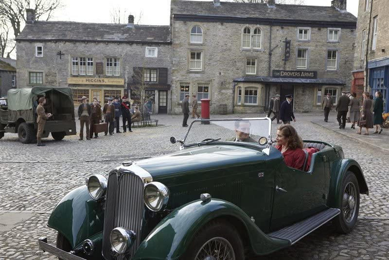 Ein grünes Oldtimer-Auto steht auf einem kopfgepflasterten Marktplatz vor historischen Steingebäuden und Passanten.
