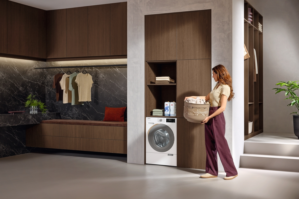 Eine Frau hält einen Wäschekorb neben einer Waschmaschine und braunen Holzschränken in einem modernen Raum.