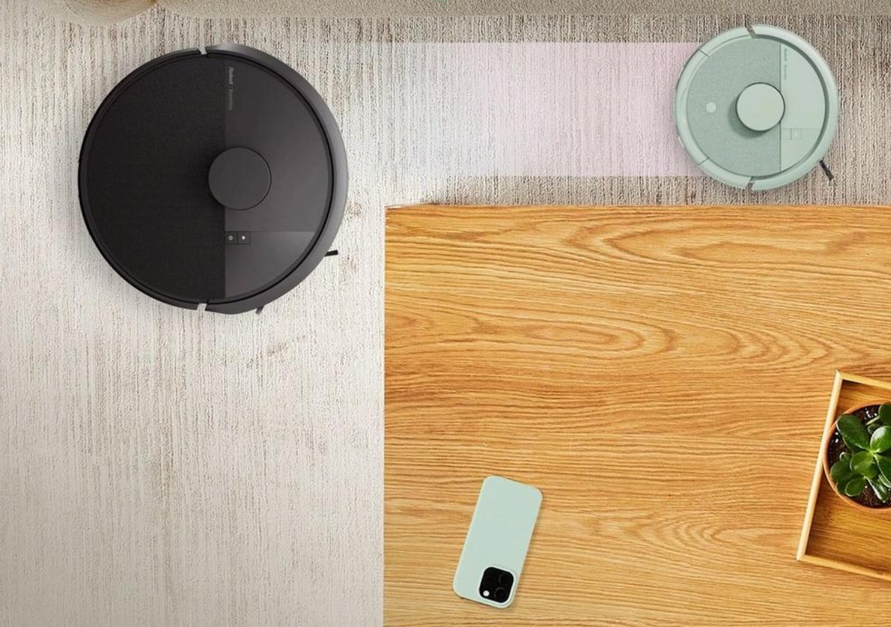 Un robot aspirador negro y otro verde en una alfombra, junto a una mesa de madera con móvil y planta.