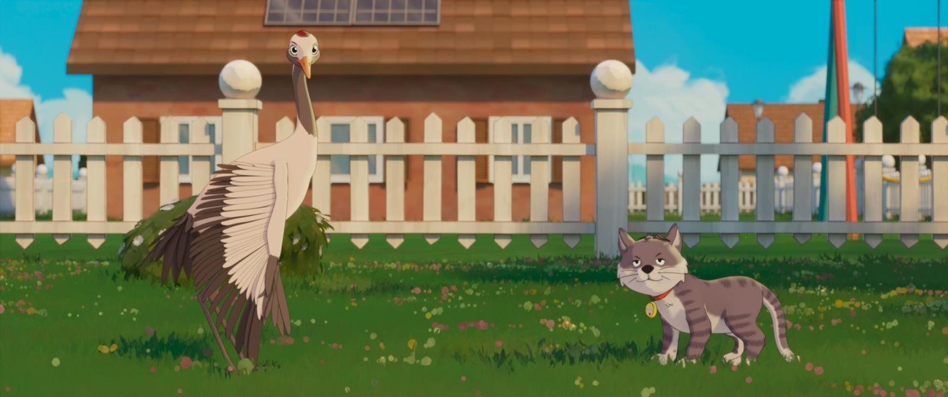 Ein Kranich und eine graue getigerte Katze stehen auf einer Wiese vor einem weißen Zaun und einem Haus.