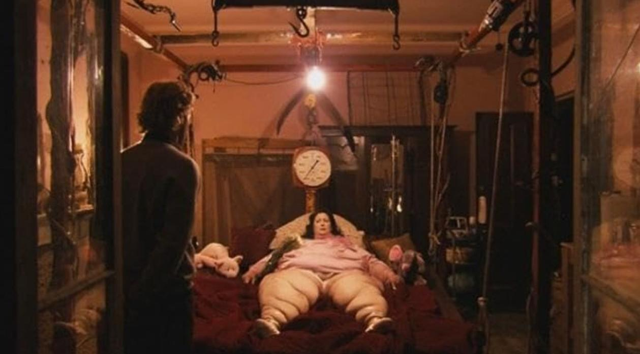 Ein Mann steht im Vordergrund, während eine übergewichtige Frau im Bett in einem dunklen, clutterhaften Raum liegt.