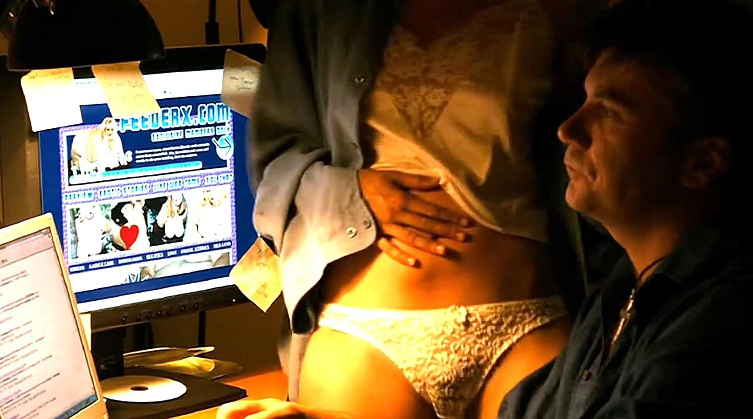 Ein Mann sitzt an einem Computer, während eine Frau in Unterwäsche und offenem Hemd vor ihm steht.