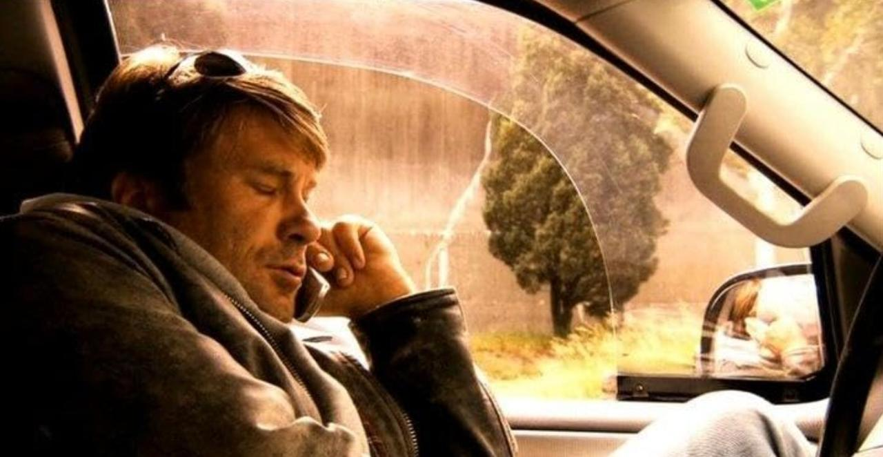 Ein Mann sitzt auf dem Beifahrersitz eines Autos und telefoniert, während draußen eine grüne Landschaft vorbeizieht.