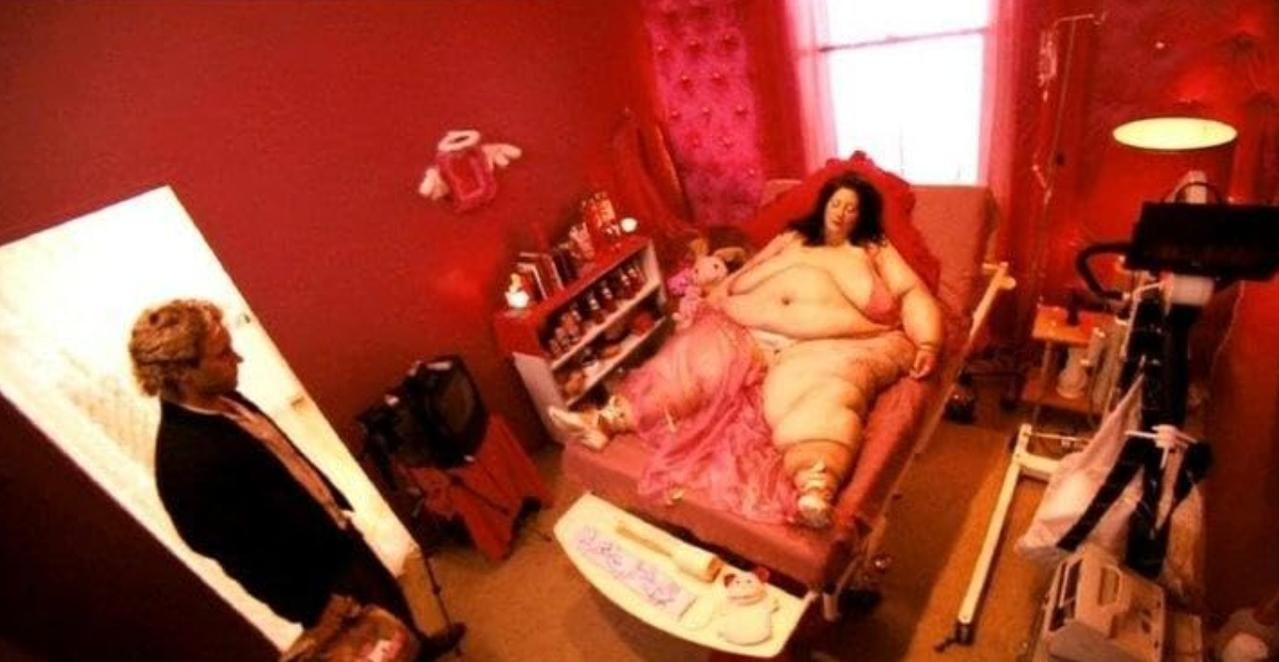 Ein Mann im Anzug steht in einem rot getönten Raum und schaut auf eine schwere Frau in einem medizinischen Bett.