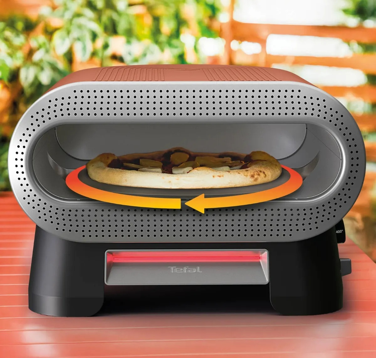 Srebrny piec do pizzy Tefal z pizzą na obracającym się kamieniu stoi na czerwonym stole.