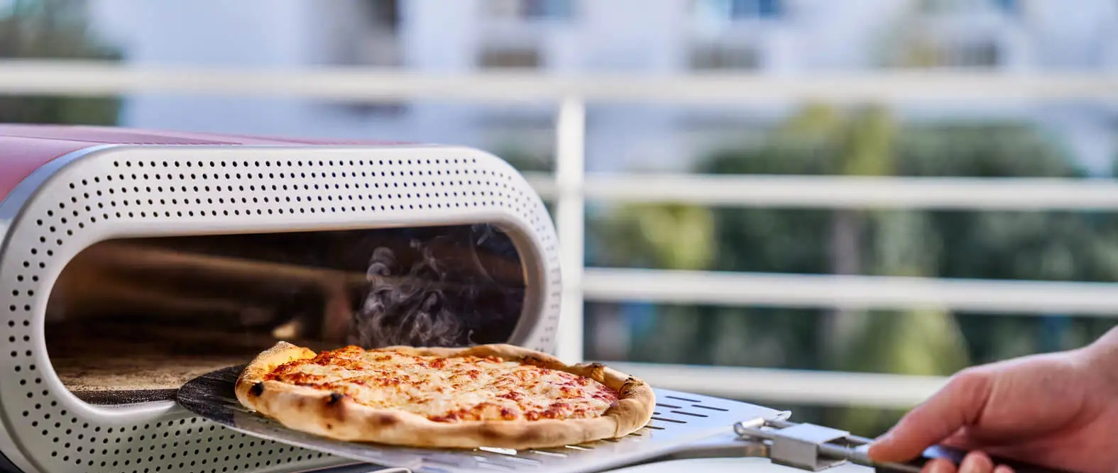 Gorąca pizza na łopacie jest wyciągana z nowoczesnego pieca zewnętrznego, unoszą się z niej opary.