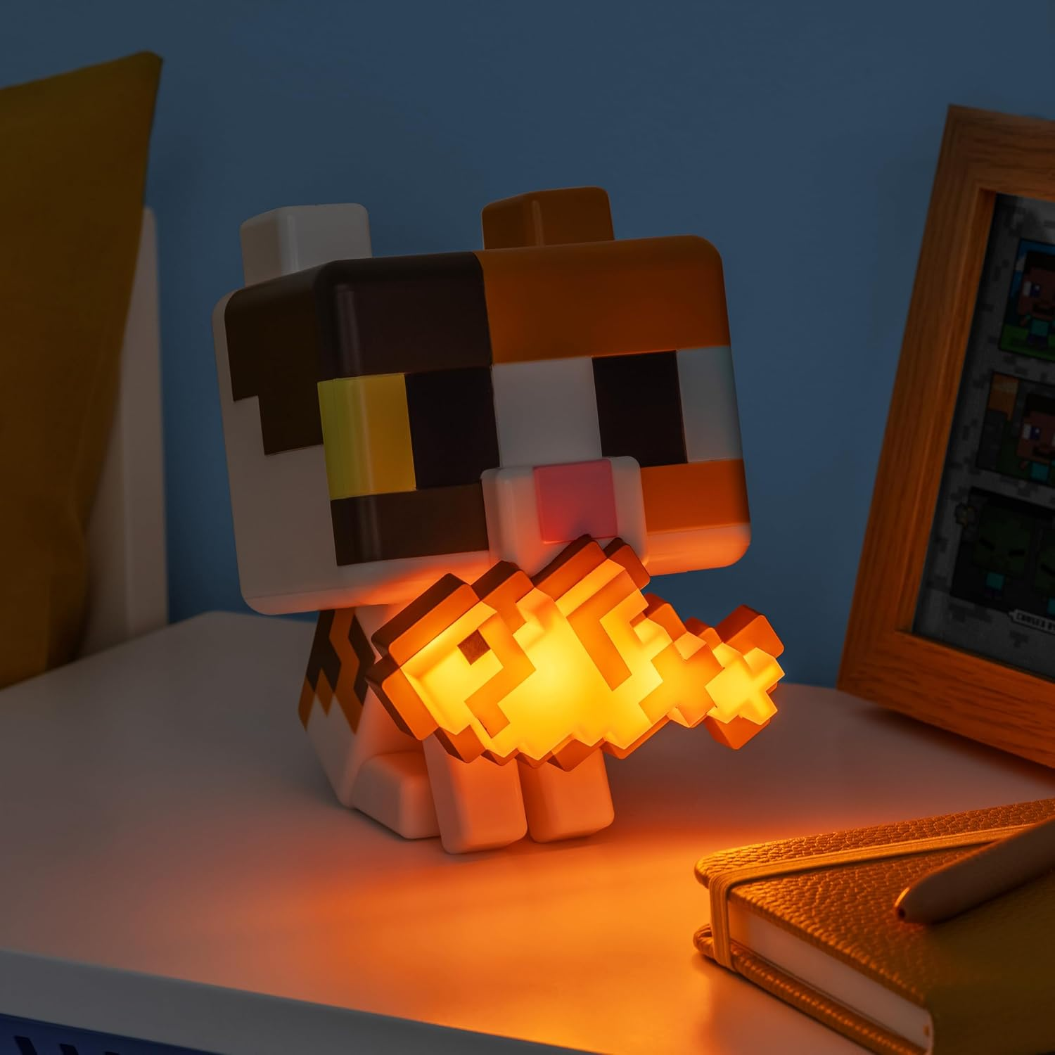Egy kockás, pixel stílusú macskalámpa narancssárga halat tart és világít egy fehér asztalon.