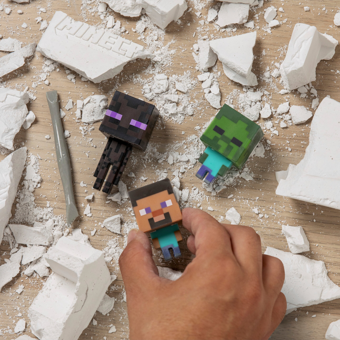 Egy kéz Minecraft figurát tart fehér gipszdarabok között, mellette Enderman, Zombi és szerszám.
