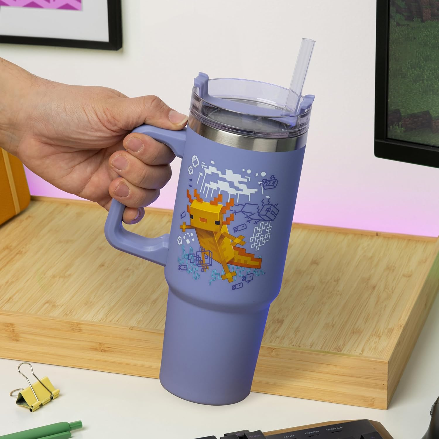 Egy kéz egy lila utazópoharat tart Minecraft axolotl grafikával, szívószállal és fa háttérrel.