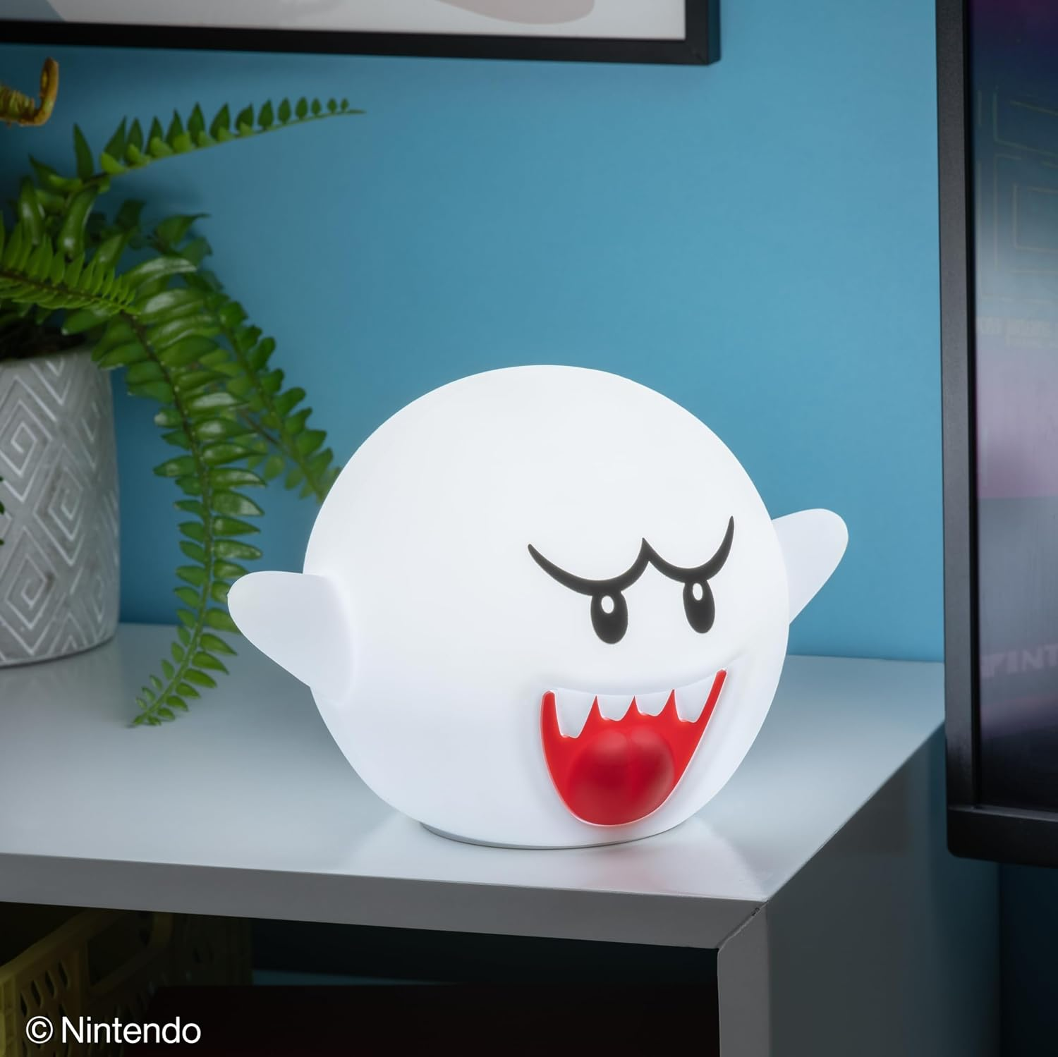 Egy fehér Super Mario Boo szellem lámpa áll az asztalon a kék fal előtt, egy növény mellett.