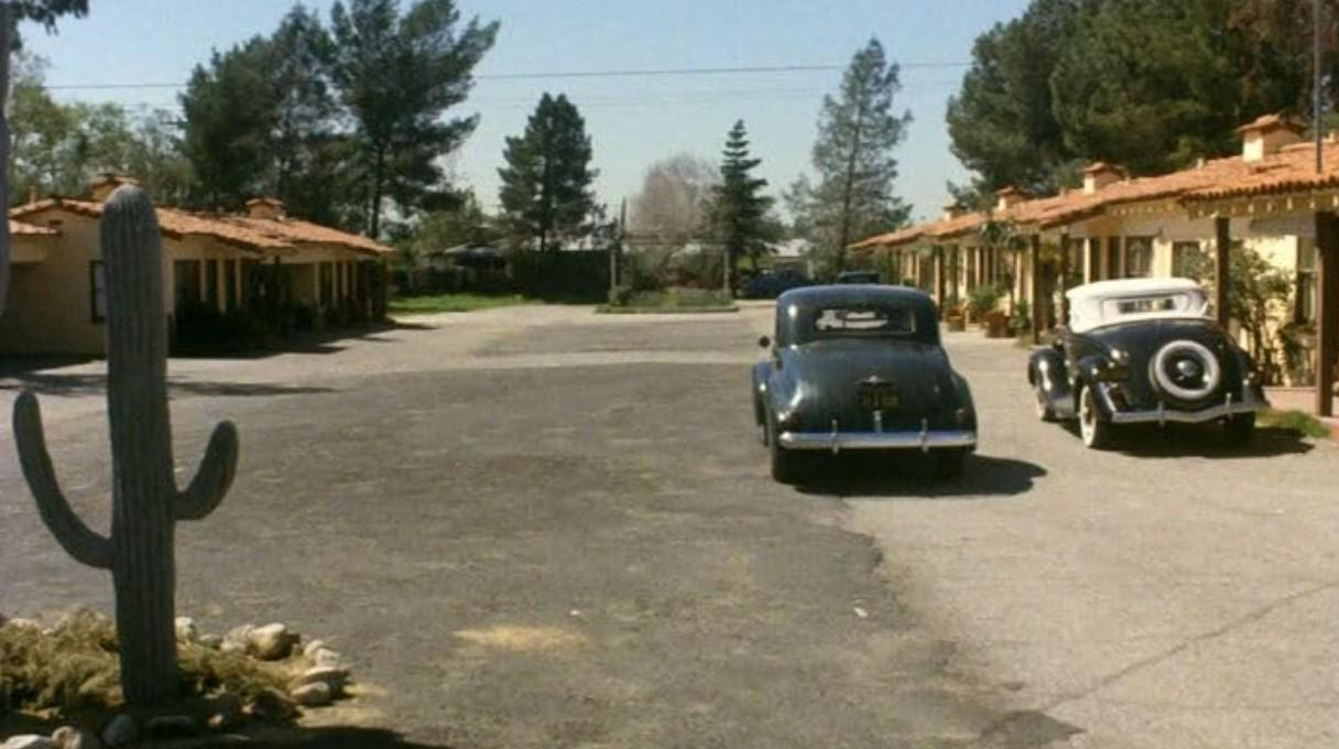 Zwei Oldtimer vor einstöckigen Motelgebäuden mit Terrakotta-Dächern und einem Kaktus im Vordergrund.