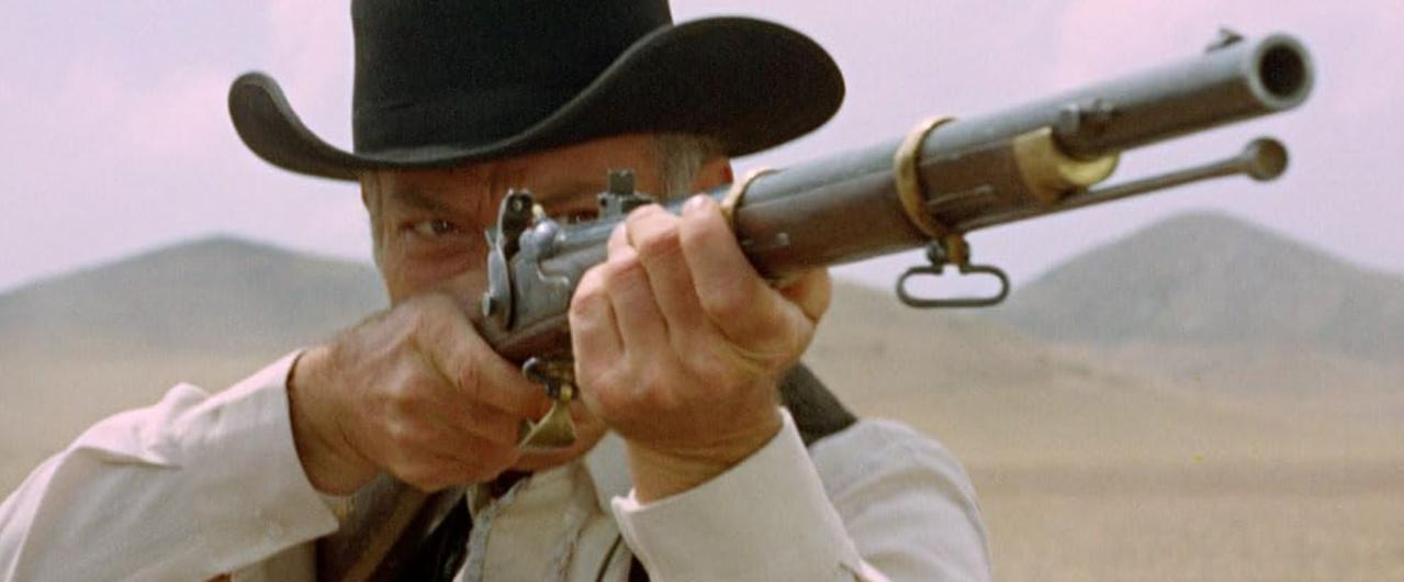Ein Cowboy mit schwarzem Hut zielt mit einem Gewehr in einer kargen Landschaft.