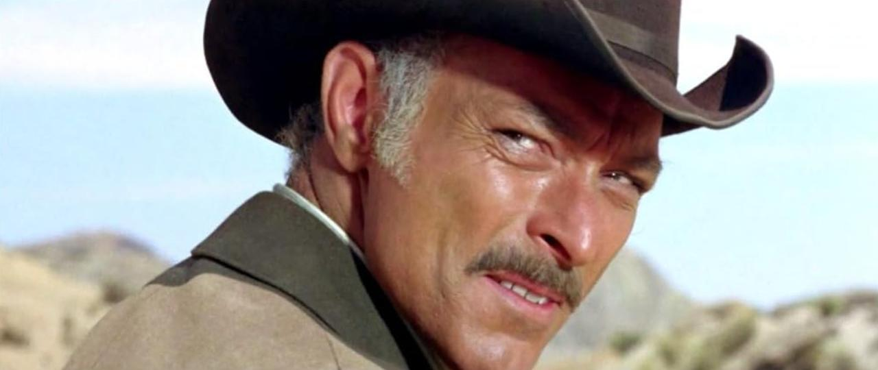 Nahaufnahme eines Mannes mit Cowboyhut und Schnurrbart vor einer kargen Landschaft.