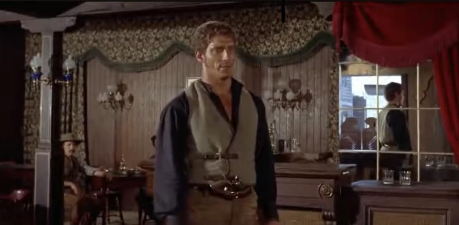 Ein Mann im Westen-Outfit steht in einem Saloon, im Hintergrund Holzwände und Spiegel.