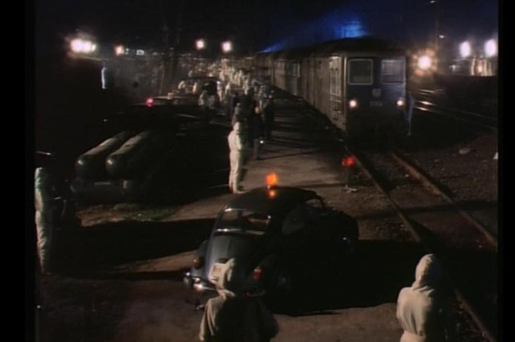 Nachtszenario an einem Bahnsteig mit einem Zug, mehreren Autos und Menschen in Schutzkleidung unter Beleuchtung.