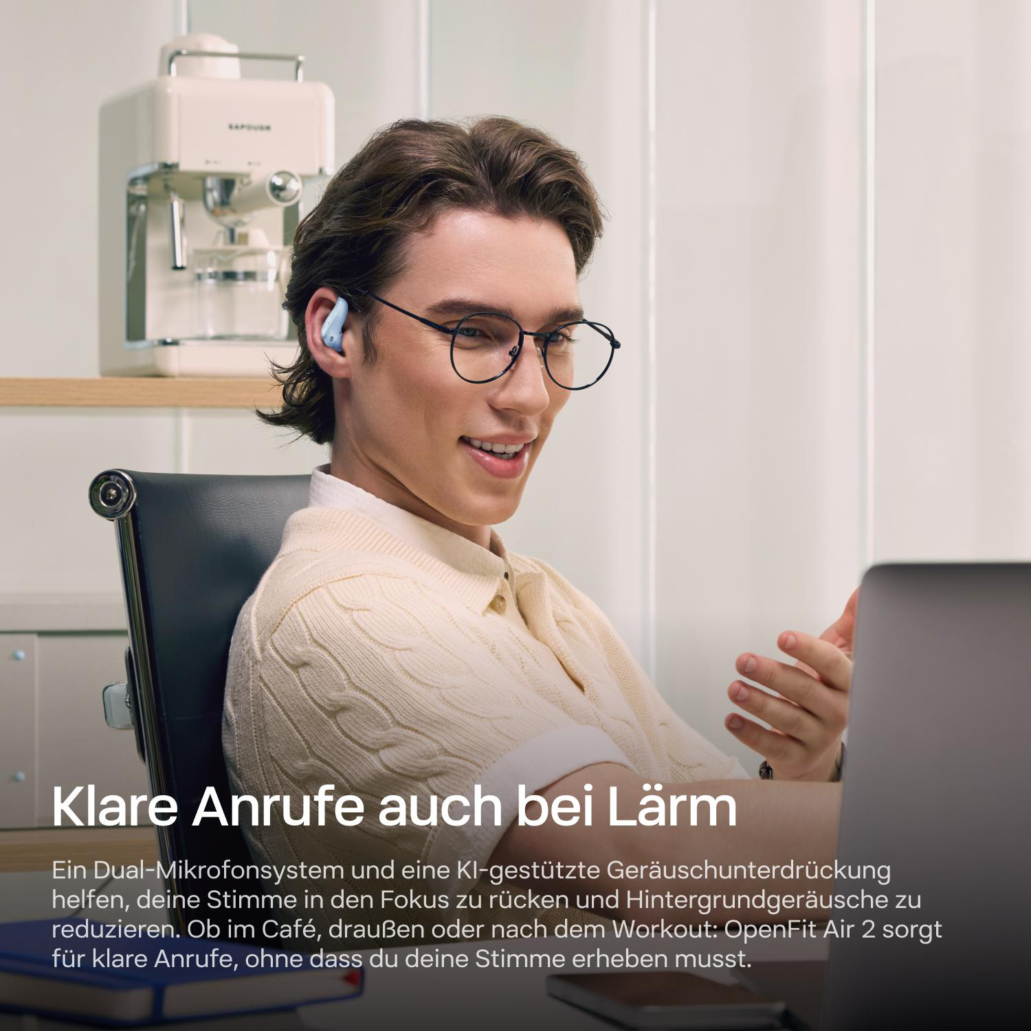 Ein Mann mit Brille und blauen Ohrhörern sitzt an einem Laptop in einem Raum mit Kaffeemaschine im Hintergrund.