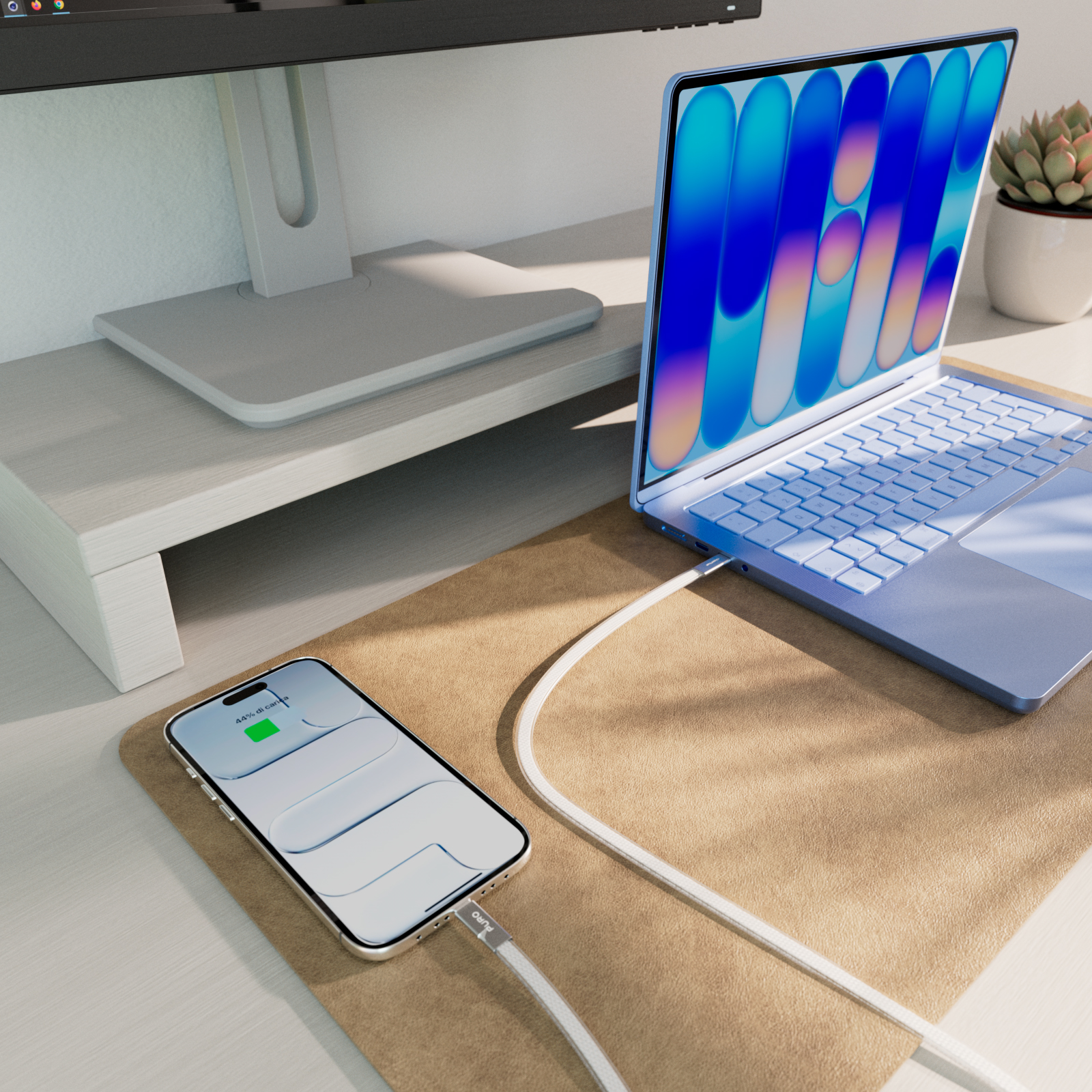 Ein Smartphone lädt über ein Kabel an einem blauen Laptop auf einer Schreibtischunterlage, mit Monitor im Hintergrund.