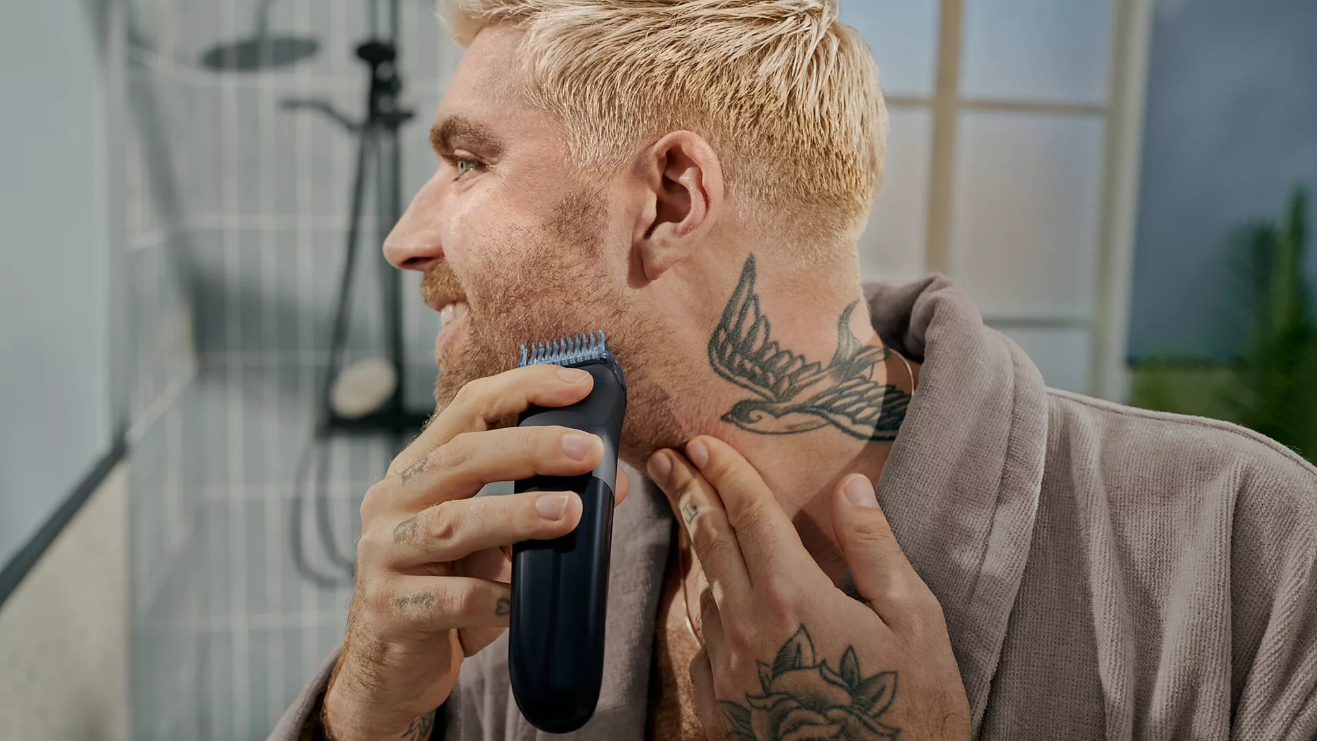 Ein blondierter Mann mit Tattoos rasiert sich im Badezimmer den Bart mit einem elektrischen Rasierer.