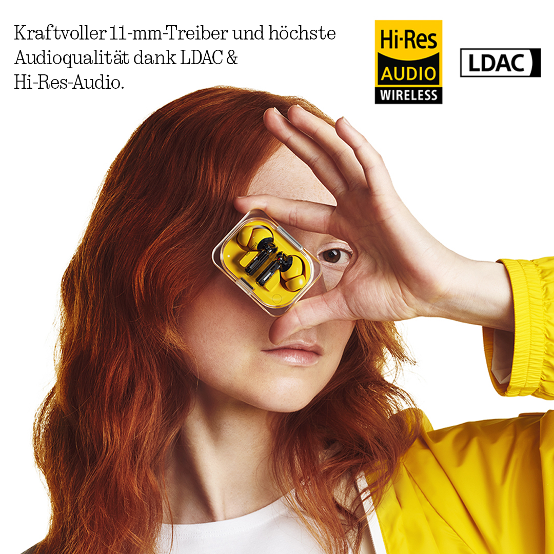 Eine rothaarige Frau in gelber Jacke hält ein durchsichtiges Etui mit gelben Ohrhörern vor das Auge.