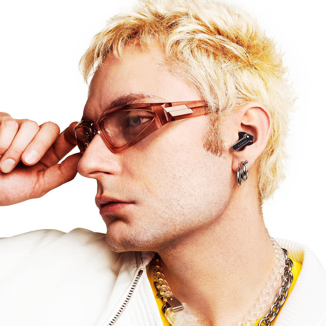 Blonder Mann mit rosafarbenen Sonnenbrillen, Ohrringen, Halsketten und drahtlosen Ohrhörern.