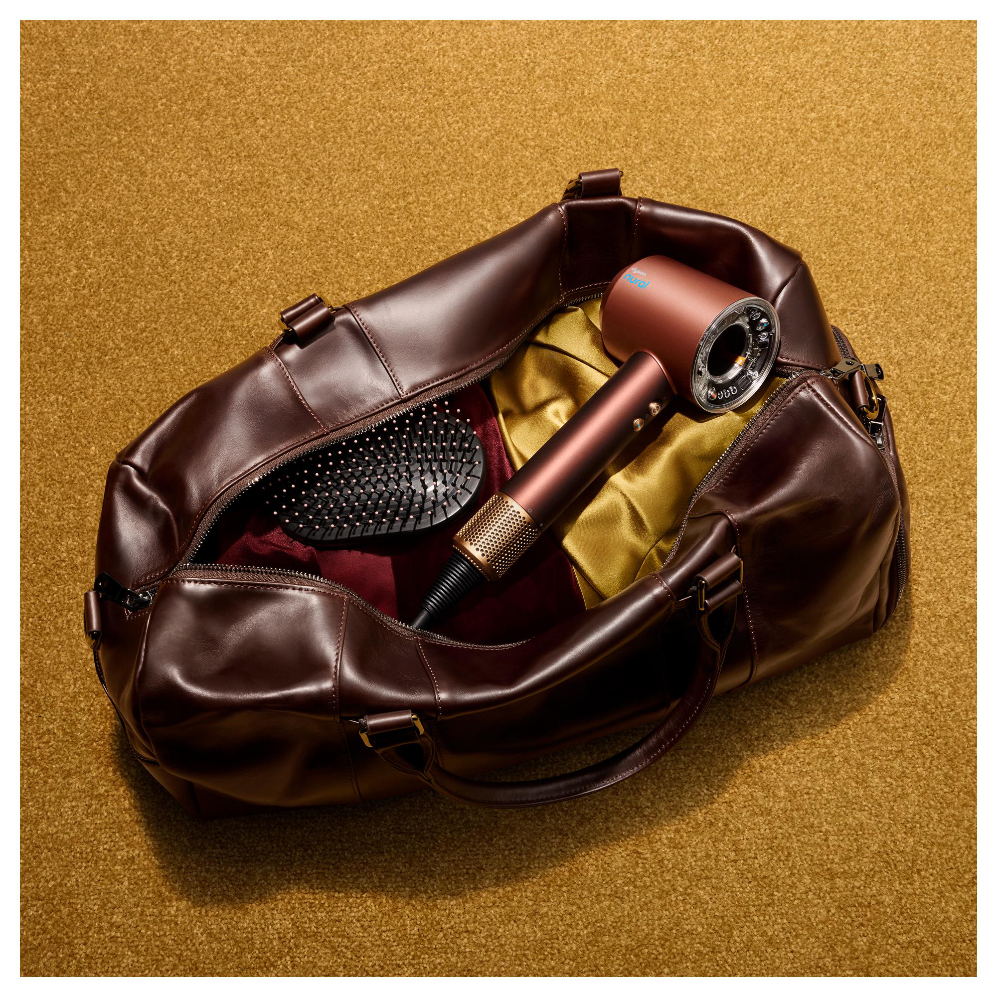 Eine braune Lederreisetasche auf gelbem Teppich enthält einen Haartrockner und eine Haarbürste.