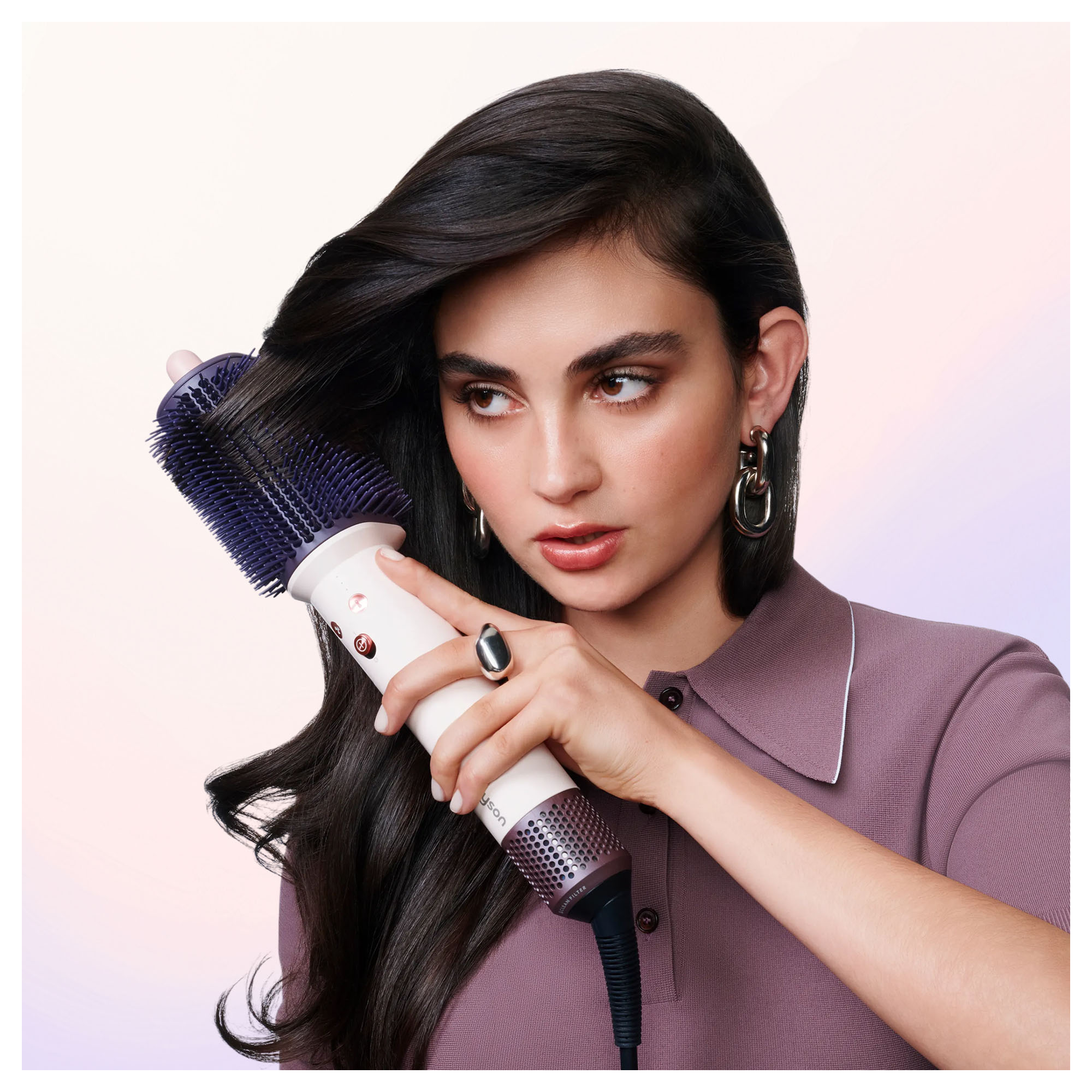 Eine Frau mit langem dunklen Haar stylt ihr Haar mit einer großen runden Haarbürste vor einem hellen Hintergrund.