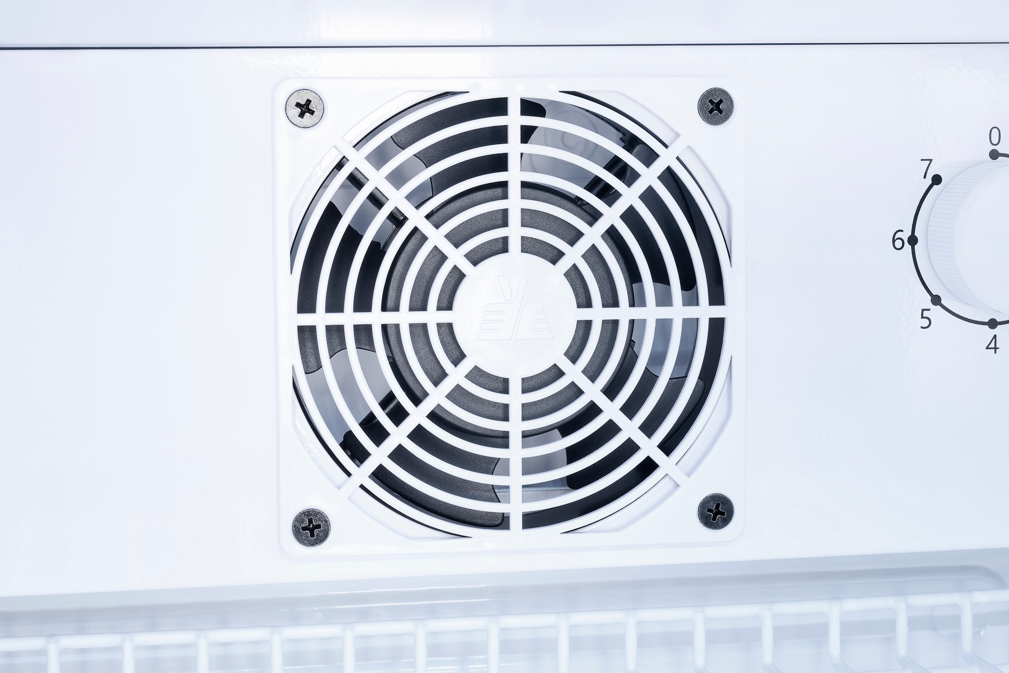 Ein weißer Lüfter mit Gitterschutz ist in eine weiße Wand mit vier Schrauben montiert, rechts ein Temperaturregler.