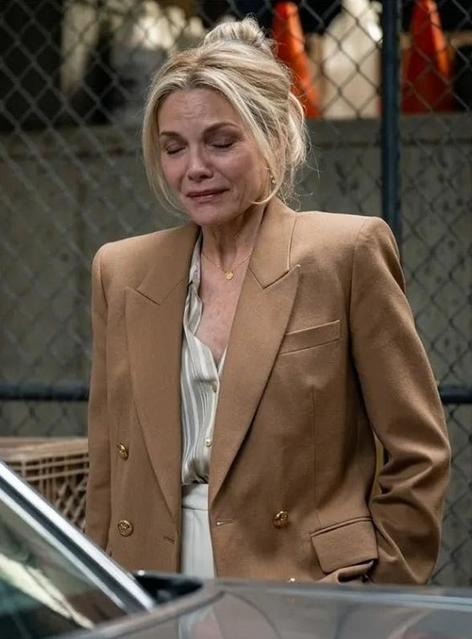 Eine blonde Frau im braunen Blazer weint mit geschlossenen Augen vor einem Drahtzaun.