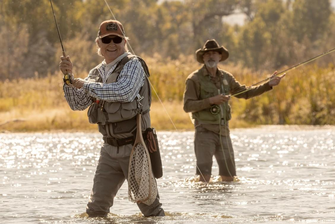Zwei Männer beim Fliegenfischen in einem sonnigen Fluss, mit Westen, Hüten und Angelruten.