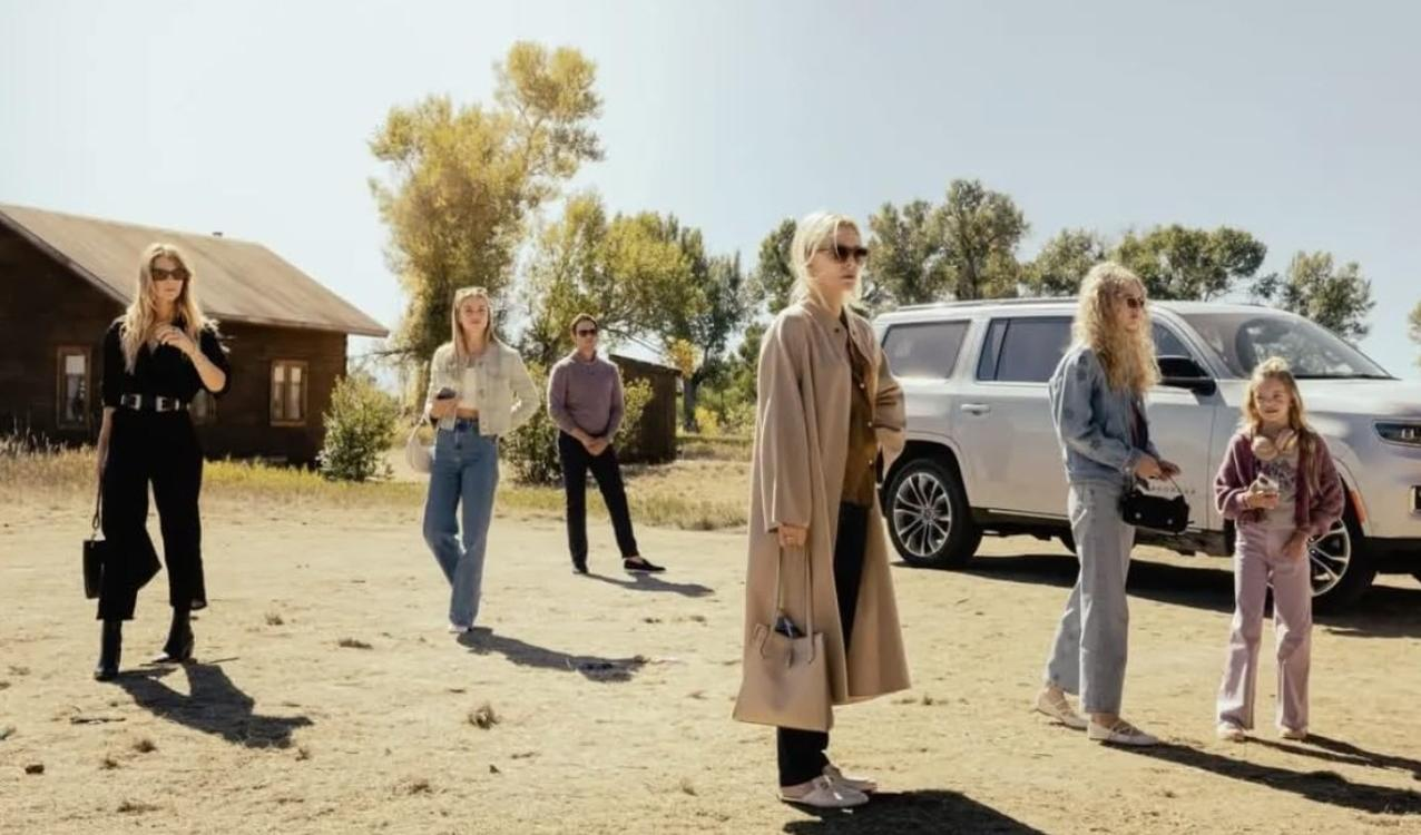Sechs Personen stehen im Freien in einer ländlichen Landschaft mit einem Holzgebäude und einem weißen SUV.