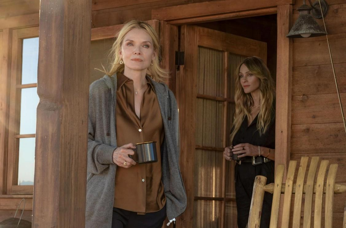 Zwei blonde Frauen stehen in der Tür einer Holzhütte, eine hält eine Tasse und die andere steht hinter ihr.