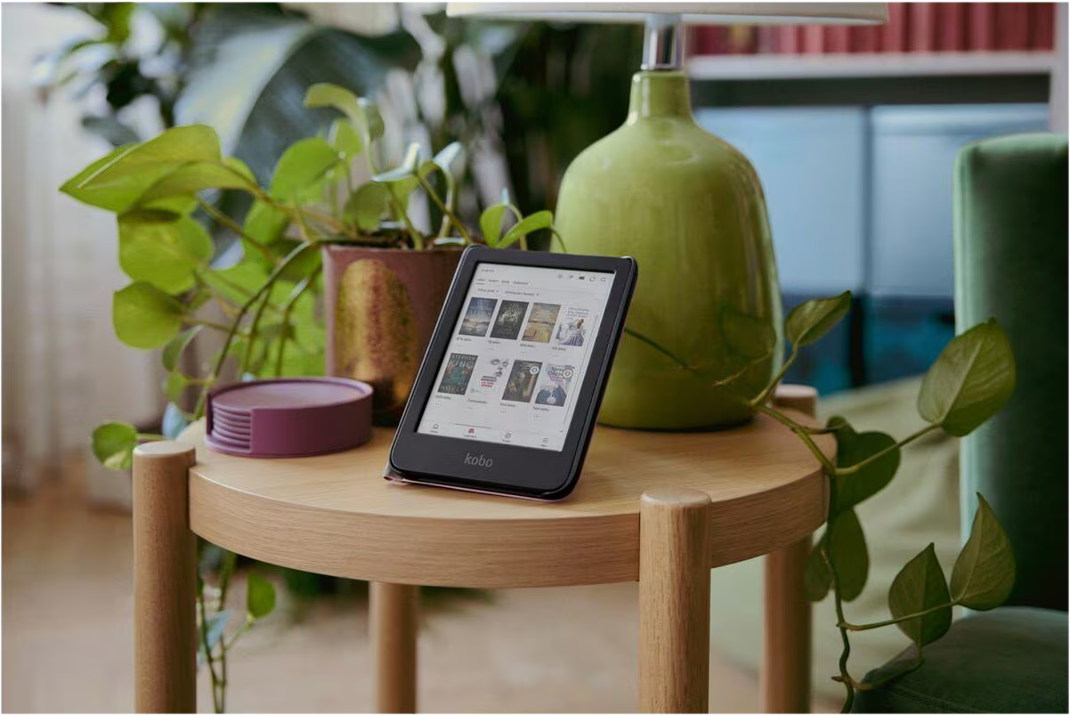 Ein E-Reader steht auf einem Holztisch, umgeben von grünen Pflanzen und einer grünen Lampe.