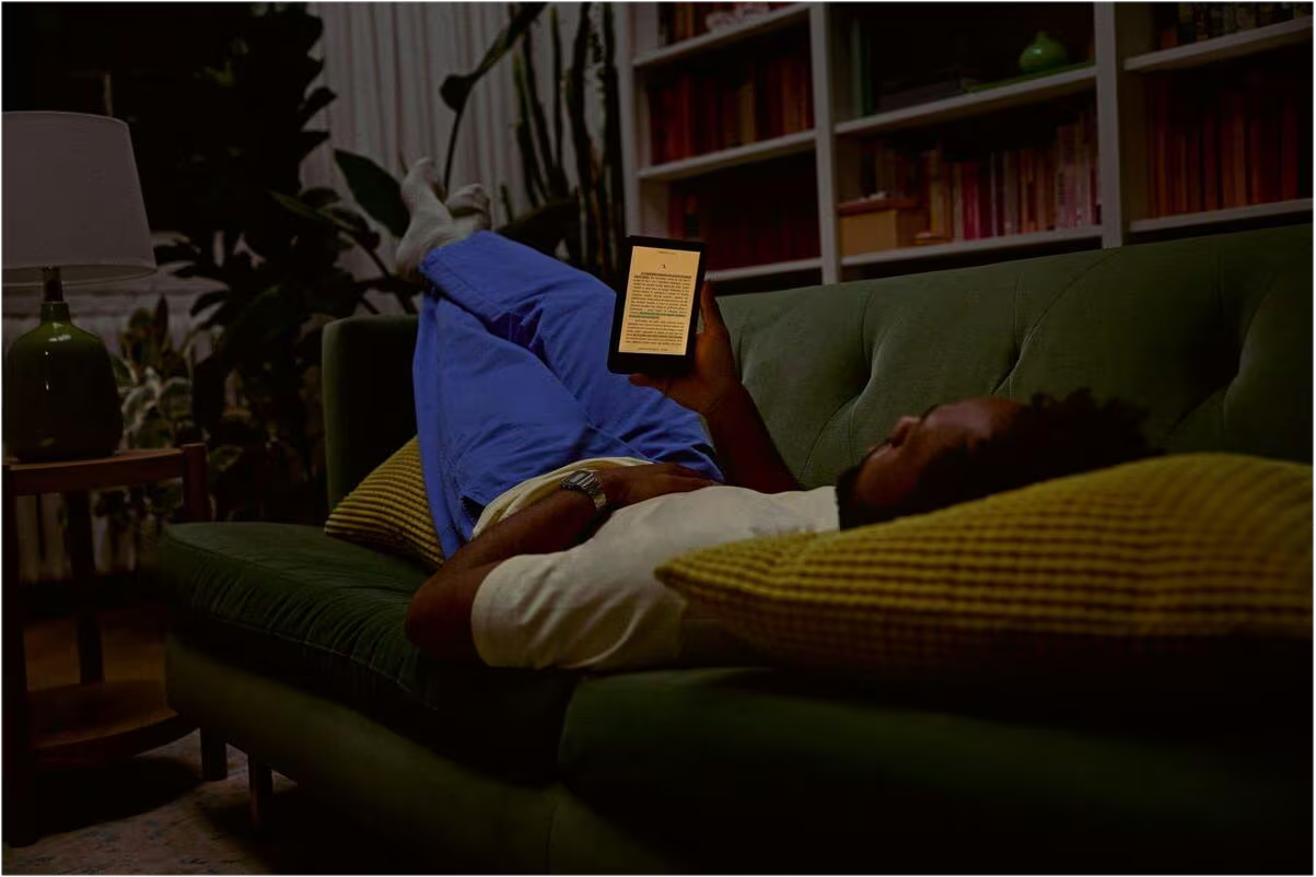 Ein Mann liegt nachts auf einem grünen Sofa in einem Wohnzimmer und liest ein E-Reader.