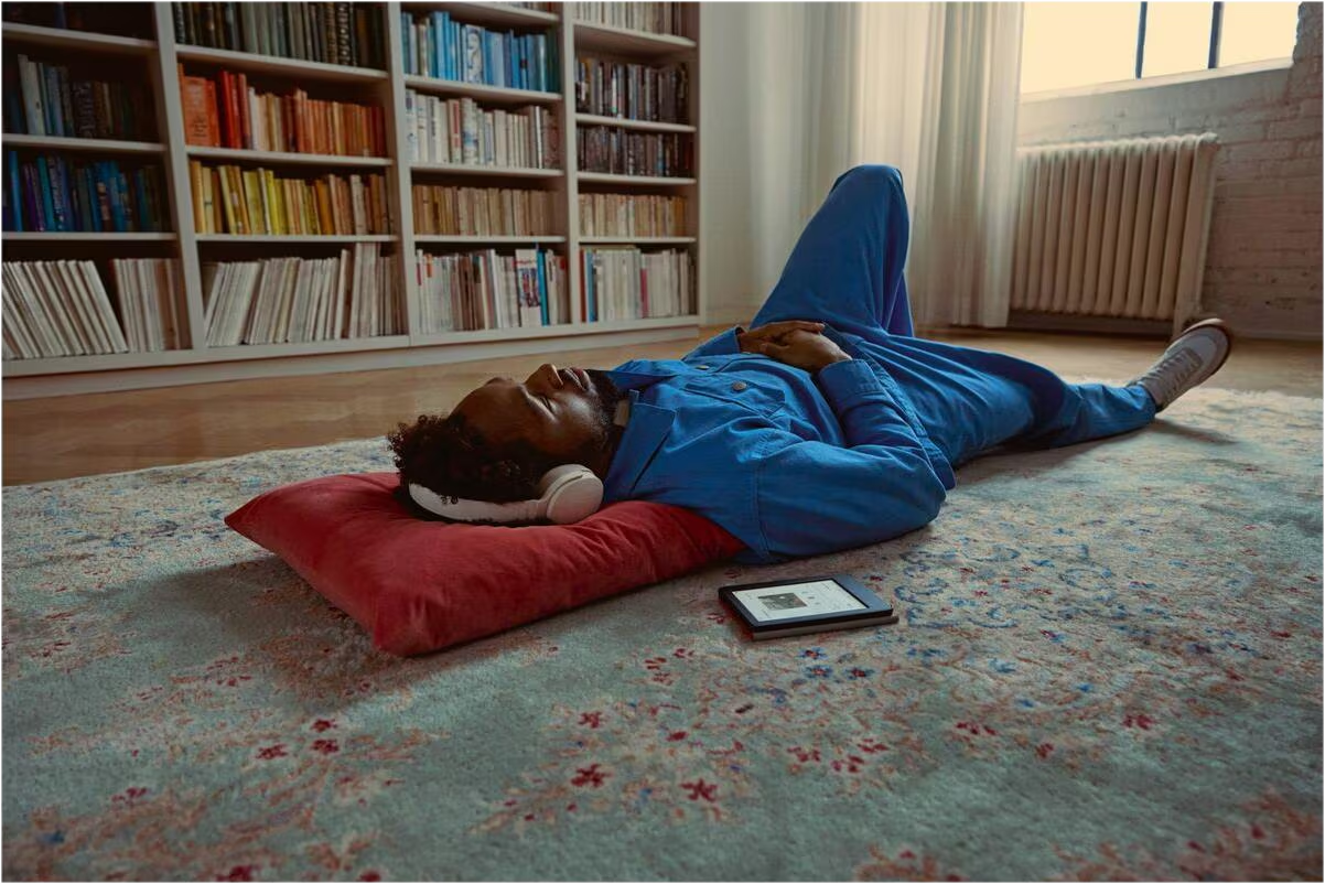 Ein Mann im blauen Overall mit Kopfhörern liegt entspannt auf einem roten Kissen auf einem Teppich vor einem großen Bücherregal.