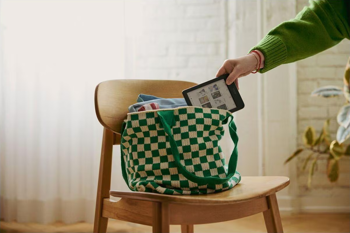 Eine Person im grünen Pullover steckt ein E-Reader-Tablet in eine karierte Tasche auf einem Holzstuhl.