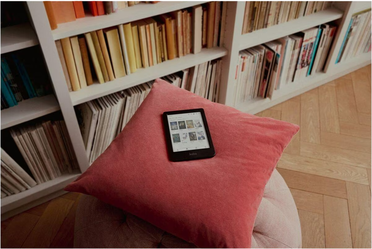 Ein schwarzer E-Reader liegt auf einem roten Kissen auf einem Hocker vor einem weißen Bücherregal.