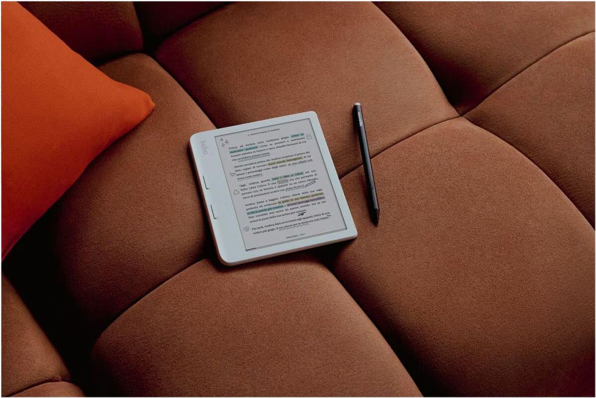 Ein weißer E-Reader und ein schwarzer Stift liegen auf einem braunen Sofa neben einem orangen Kissen.
