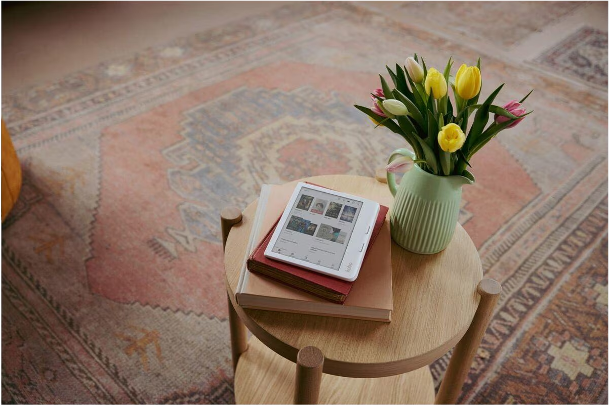Ein E-Reader auf Büchern und eine Vase mit gelben und rosa Tulpen auf einem kleinen Holztisch über einem Teppich.