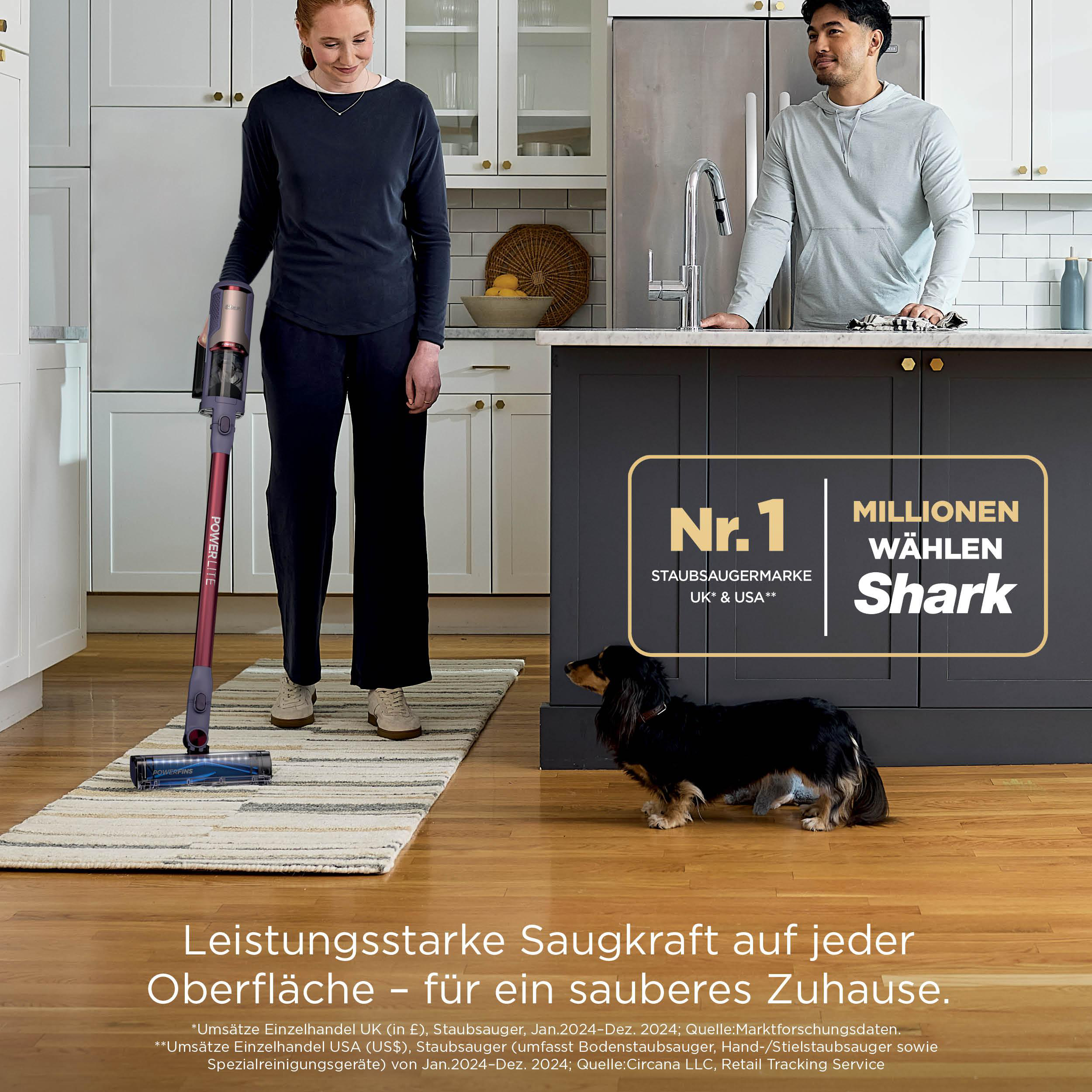 Eine Frau saugt einen Teppich mit einem kabellosen Shark-Staubsauger, während ein Mann und ein Hund zusehen.