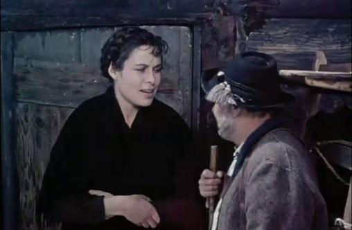Eine Frau in Schwarz spricht in einer Holzhütte mit einem Mann im Hut mit Stock.