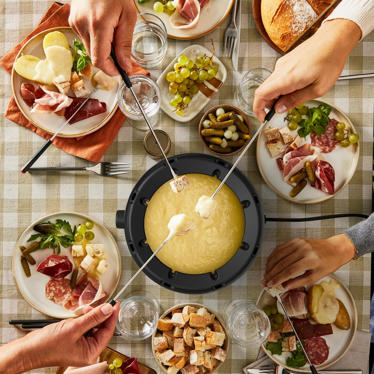 Four people enjoying cheese fondue at a table with bread, potatoes, ham, and pickles.